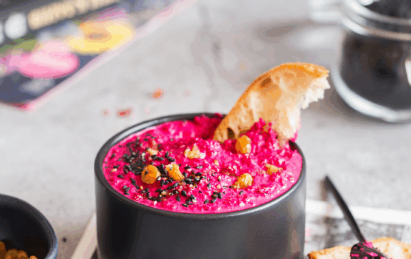 Petit bol noir remplie de tapenade de betterave et noix, parsemé de graine de sésame avec une tranche de pain sur un morceau de journal
